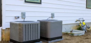 Two gray residential air conditioning condenser units installed outside a white house with metal piping and electrical connections visible.