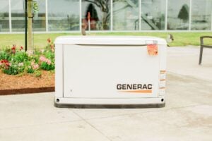 White Generac standby generator unit installed on concrete pad beside landscaped flower beds with greenhouse visible in background.