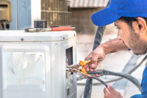 Technician in blue cap using pliers to repair electrical wiring connections on white outdoor air conditioning unit.