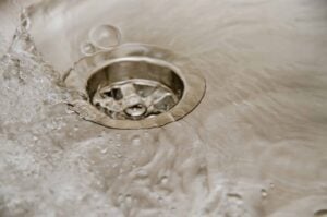 Water flowing down a residential sink drain with splashing water droplets visible around the metal drain opening.